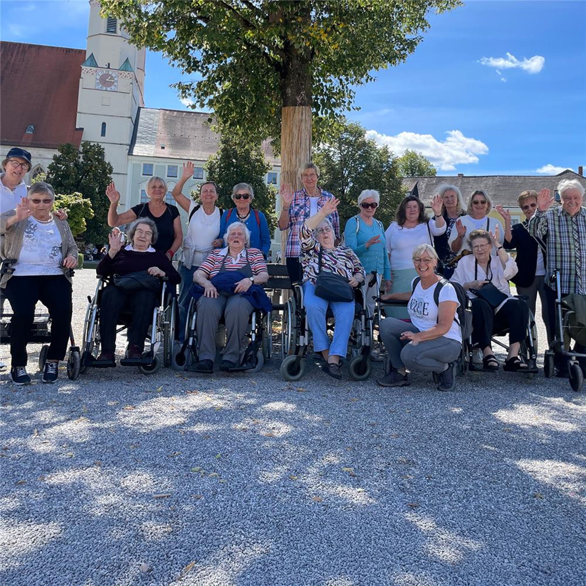 Eine Gruppe von Menschen steht im freien auf einem Platz mit Kieselsteinen. Im Hintergrund ist ein Baum und eine Kirche.